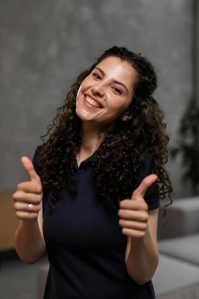 Cheerful woman with curly hair giving thumbs up indoors, smiling brightly.